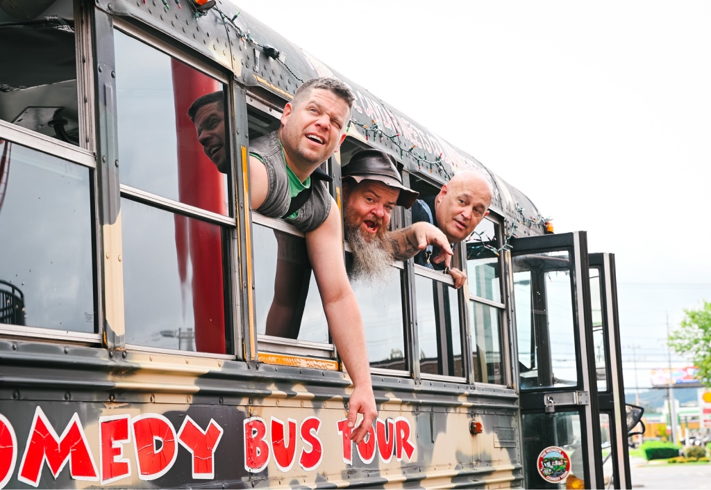 Three men lean out of a camouflaged painted comedy tour bus with expressions of excitement and amusement.