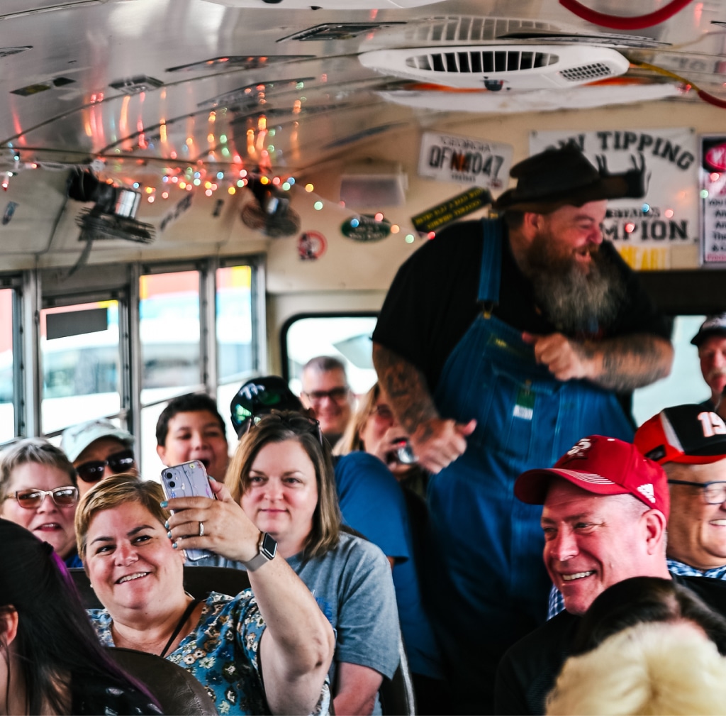 A group of people, some standing and some sitting, are enjoying themselves inside a decorated bus. One person in the foreground is taking a selfie, while another person in overalls is standing and smiling.