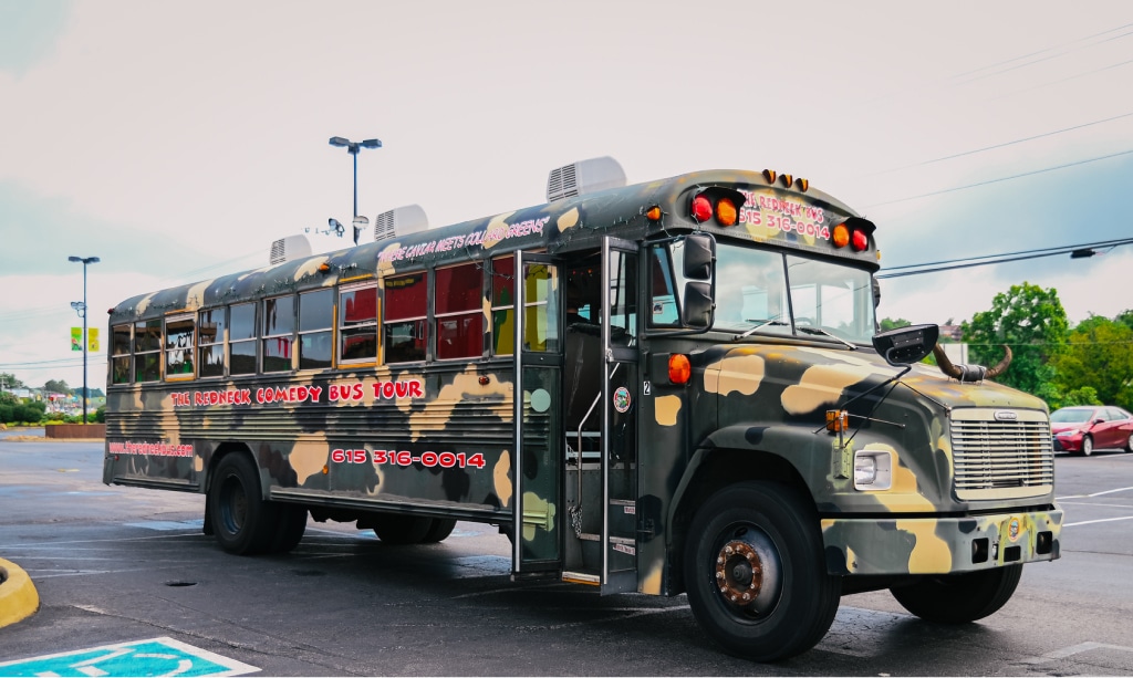 A large, camo-painted tour bus with "Redneck Comedy Bus Tour" written on the side, parked in a parking lot with blue sky and trees in the background.