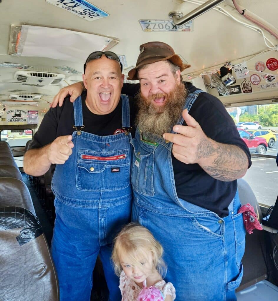 Two men in denim overalls and a young girl stand inside a bus. The men are smiling and gesturing enthusiastically, with one pointing at the camera. The girl stands in front of them, looking at the camera.