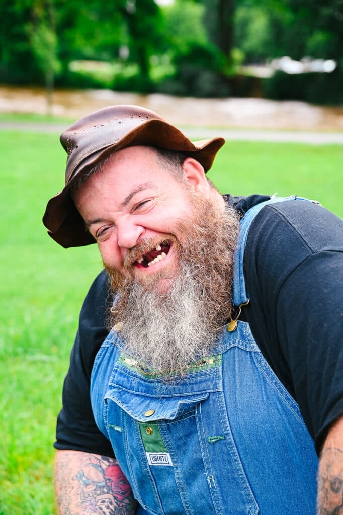 A man with a large beard and missing teeth wears a leather hat and denim overalls, smiling while sitting outside on grass.