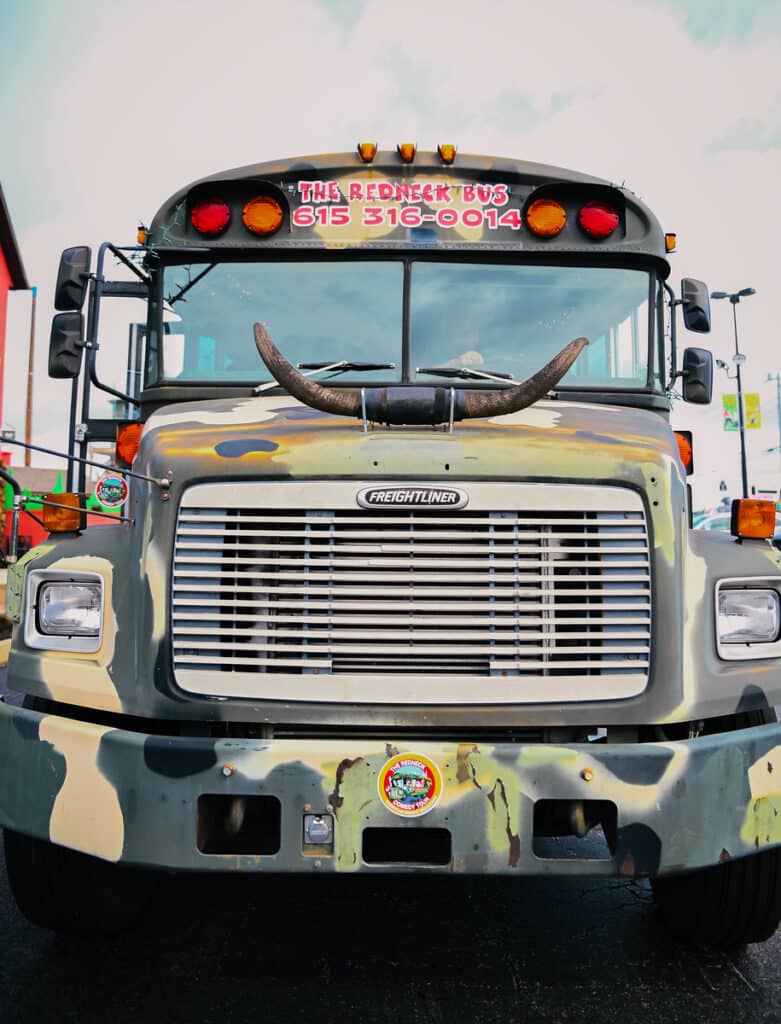 Front view of a camouflaged bus with bull horns attached to the hood and the text "The Redneck Bus" along with a phone number displayed above the windshield.