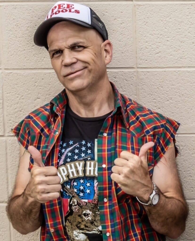 A man wearing a checkered shirt, black graphic t-shirt, and cap stands against a tiled wall. He is smiling and giving two thumbs up.