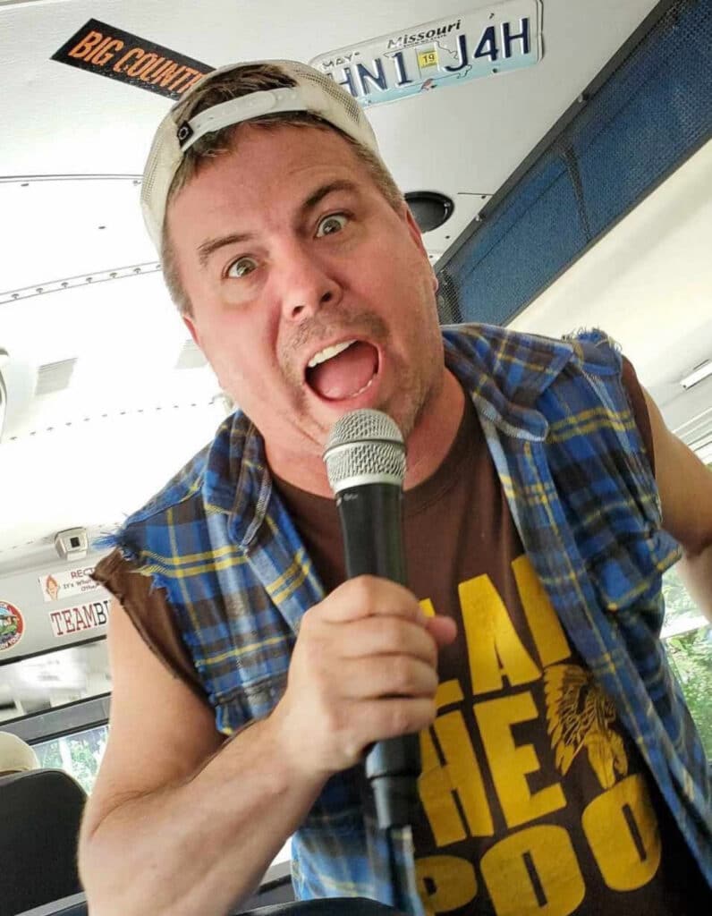 A man wearing a cap backward, a plaid vest over a brown T-shirt, and holding a microphone, appears to be passionately speaking or singing on a bus.