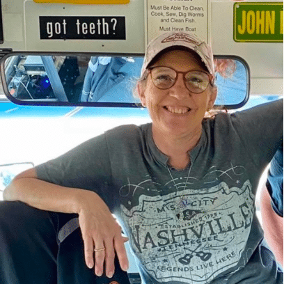 A smiling person in a 'Nashville' t-shirt and cap leans against a surface. Various humorous signs are visible in the background.