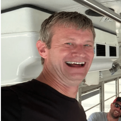 A man with short gray hair smiles while standing inside a bus, with another person slightly visible in the background.