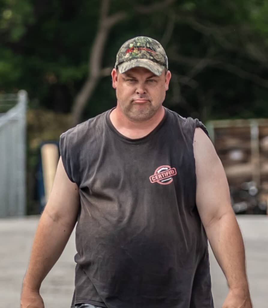 A man wearing a camouflage cap and a sleeveless shirt with "CERTIFIED" written on it, stands outdoors on a paved surface. Trees and a fence are visible in the background.