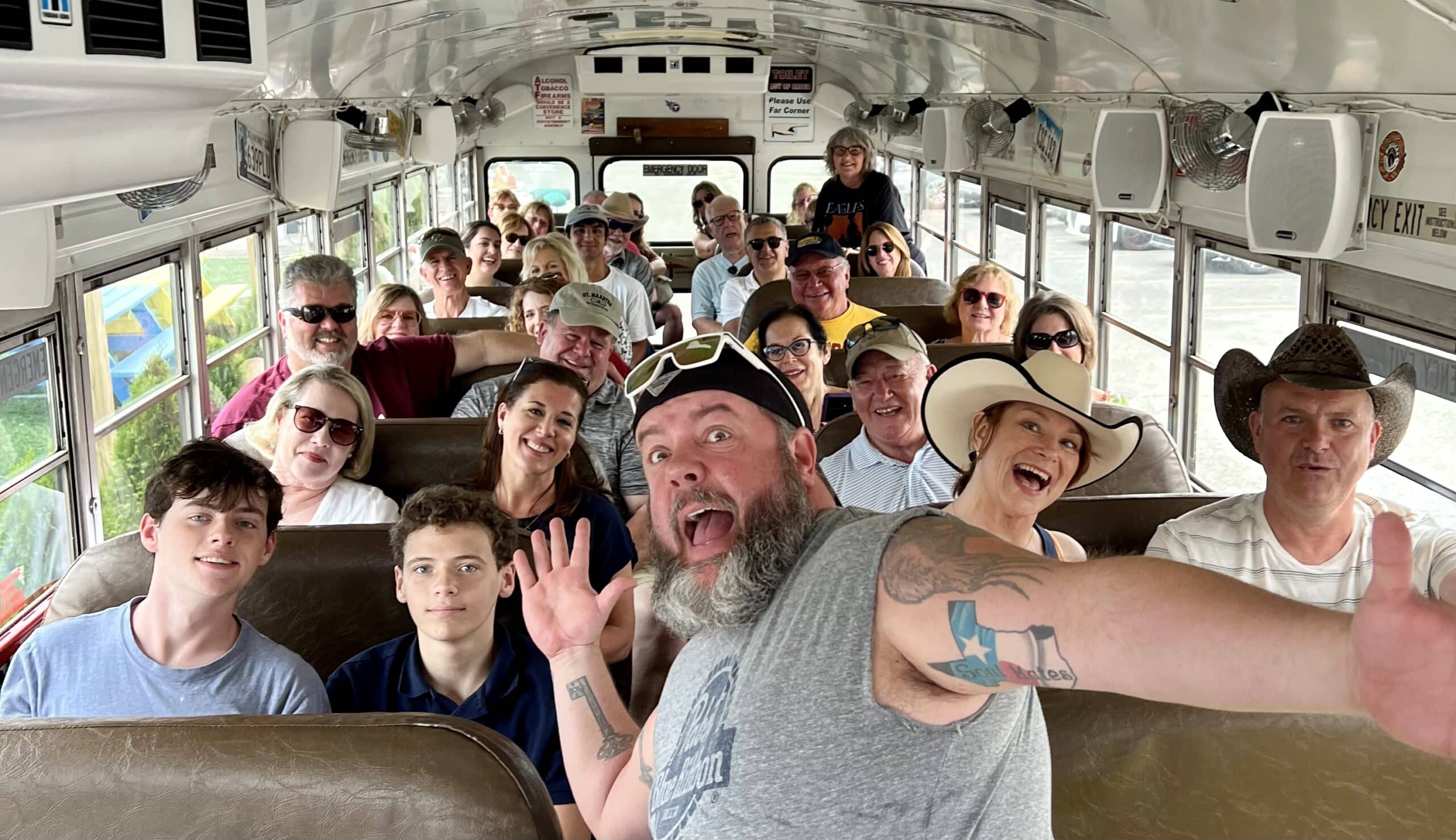 A group of people, seated inside a bus, smiling and posing for a photo. Several individuals are wearing hats and sunglasses. The bus is filled with a mix of adults and children.