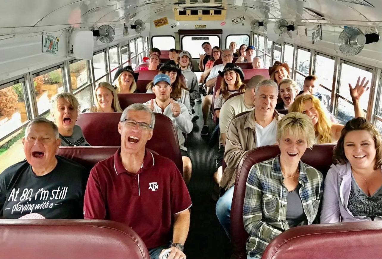A group of people, sitting and smiling on a bus, filled all the way to the back with men and women of various ages. The mood appears to be joyful and lively.