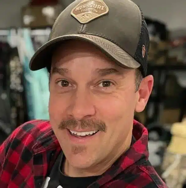 Person smiling, wearing a black and brown cap with a "Missouri Tigers" patch and a red plaid shirt. Background shows various clothing items.