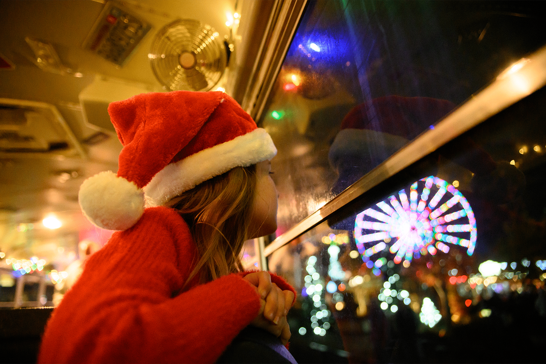 A child wearing a Santa hat looks out a bus window at a brightly lit Ferris wheel and holiday lights at night.