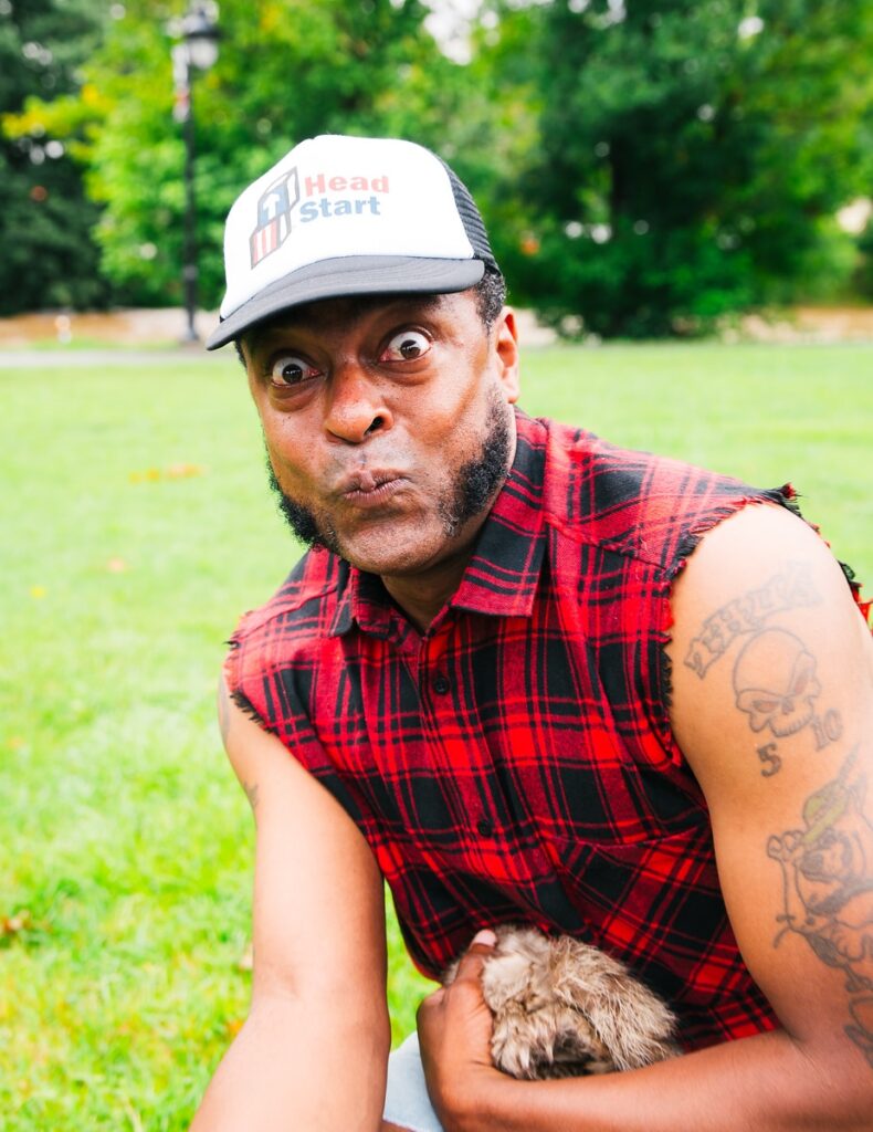 A man in a red plaid sleeveless shirt and "Head Start" cap makes a funny face while holding a small animal, sitting on grass with trees in the background.