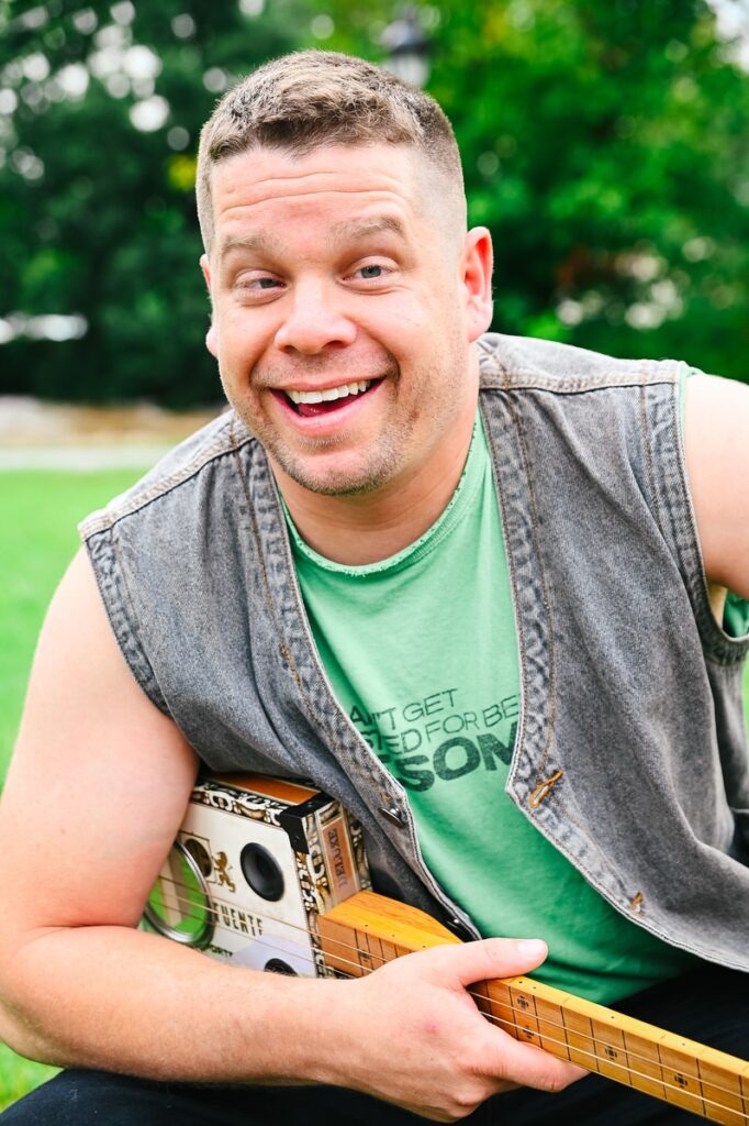 Man with short hair smiles outdoors, wearing a sleeveless denim vest and green T-shirt, holding a homemade guitar.
