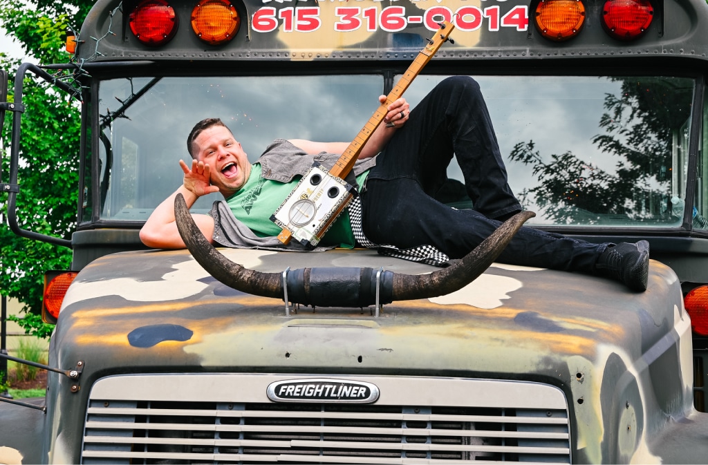 A man lies on top of a camouflaged Freightliner bus, holding a homemade guitar and making a rock gesture. The bus has large horns mounted on its front.