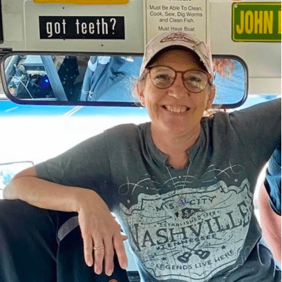 A woman wearing glasses and a cap smiles while sitting on a vehicle. Signs above her read "got teeth?" and other messages.