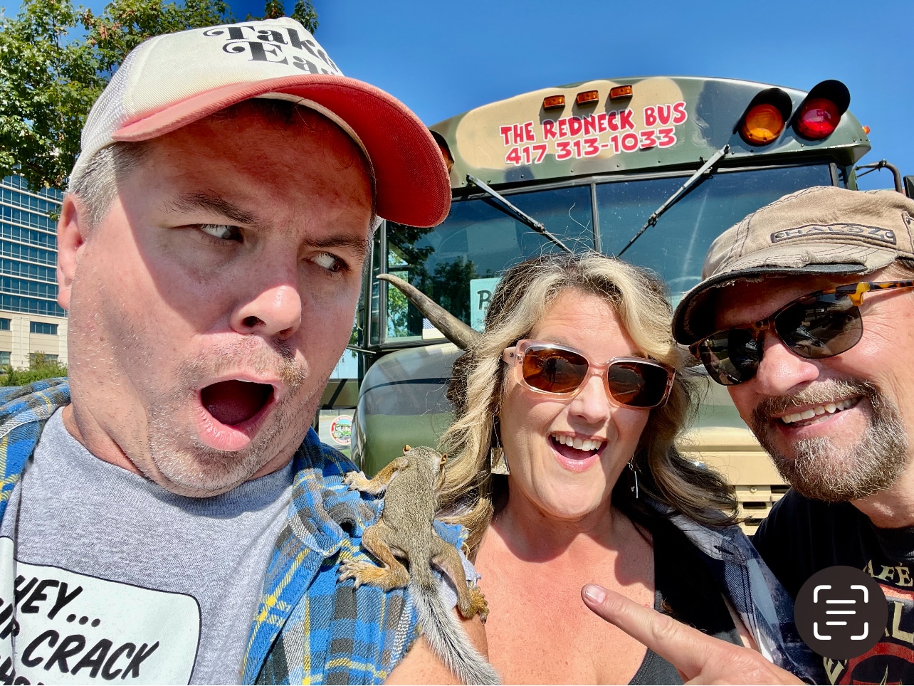 Three adults pose in front of a bus labeled "The Redneck Bus"; one man looks surprised at a squirrel perched on his shoulder.