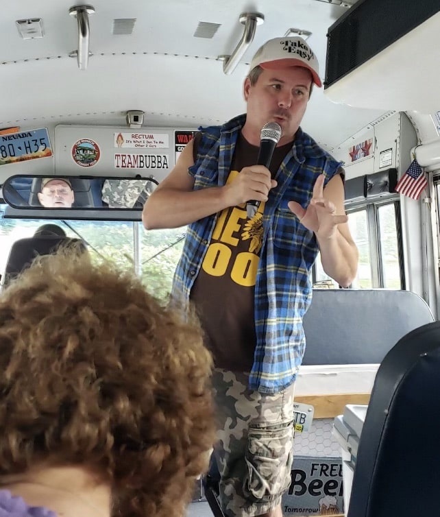 A man wearing a plaid sleeveless shirt, camouflage shorts, and a cap speaks into a microphone while standing at the front of a bus. Passengers are seated and listening.