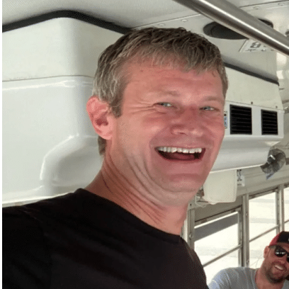 A man with short gray hair smiles broadly while standing inside a vehicle, with another person partially visible in the background.