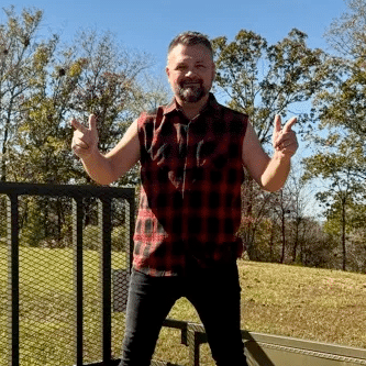 A man wearing a sleeveless plaid shirt stands outdoors on a metal structure, smiling and pointing both index fingers, with trees and grass in the background.