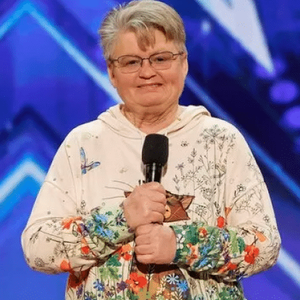 An older woman with short gray hair and glasses holds a microphone, standing on stage in front of a blue backdrop with geometric patterns.