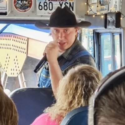 A man in a cowboy hat and overalls holds a microphone, speaking to passengers seated on a bus.