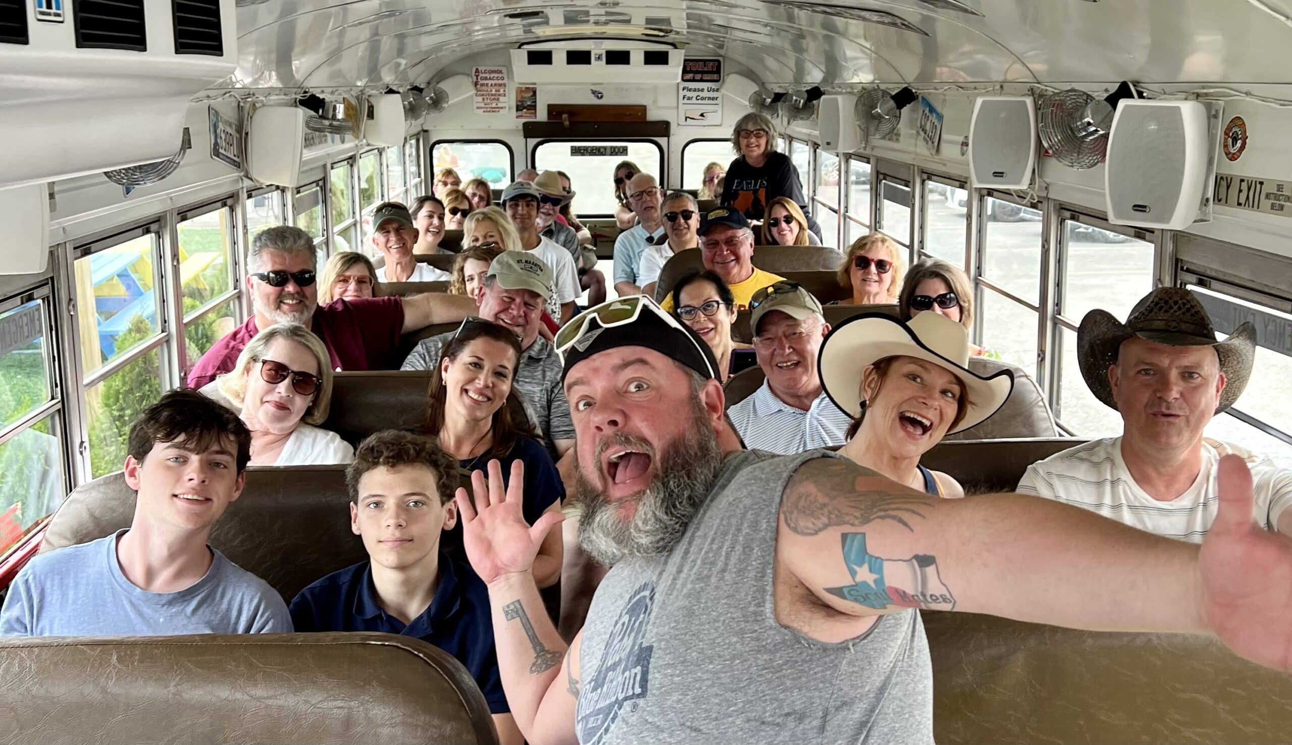 A group of adults and teens smile and pose for a selfie inside a school bus, with one man in front taking the photo and making a playful face.