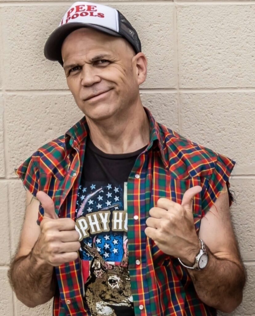 A man in a sleeveless plaid shirt, graphic tee, and cap stands against a beige wall, giving two thumbs up with a neutral facial expression.