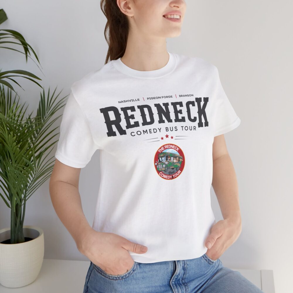 A woman indoors with hands in her jeans pockets wears the Redneck Comedy Bus Tour Tshirt. A potted plant sits in the background.