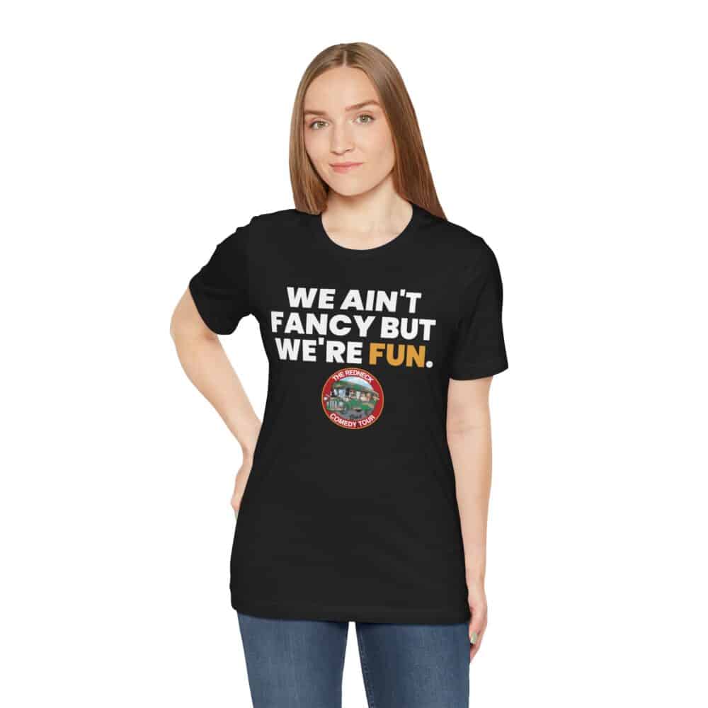 A woman poses with one hand on her hip, wearing the We Ain't Fancy Tshirt—a black top featuring "WE AIN'T FANCY BUT WE'RE FUN." and a colorful circular design below the text.