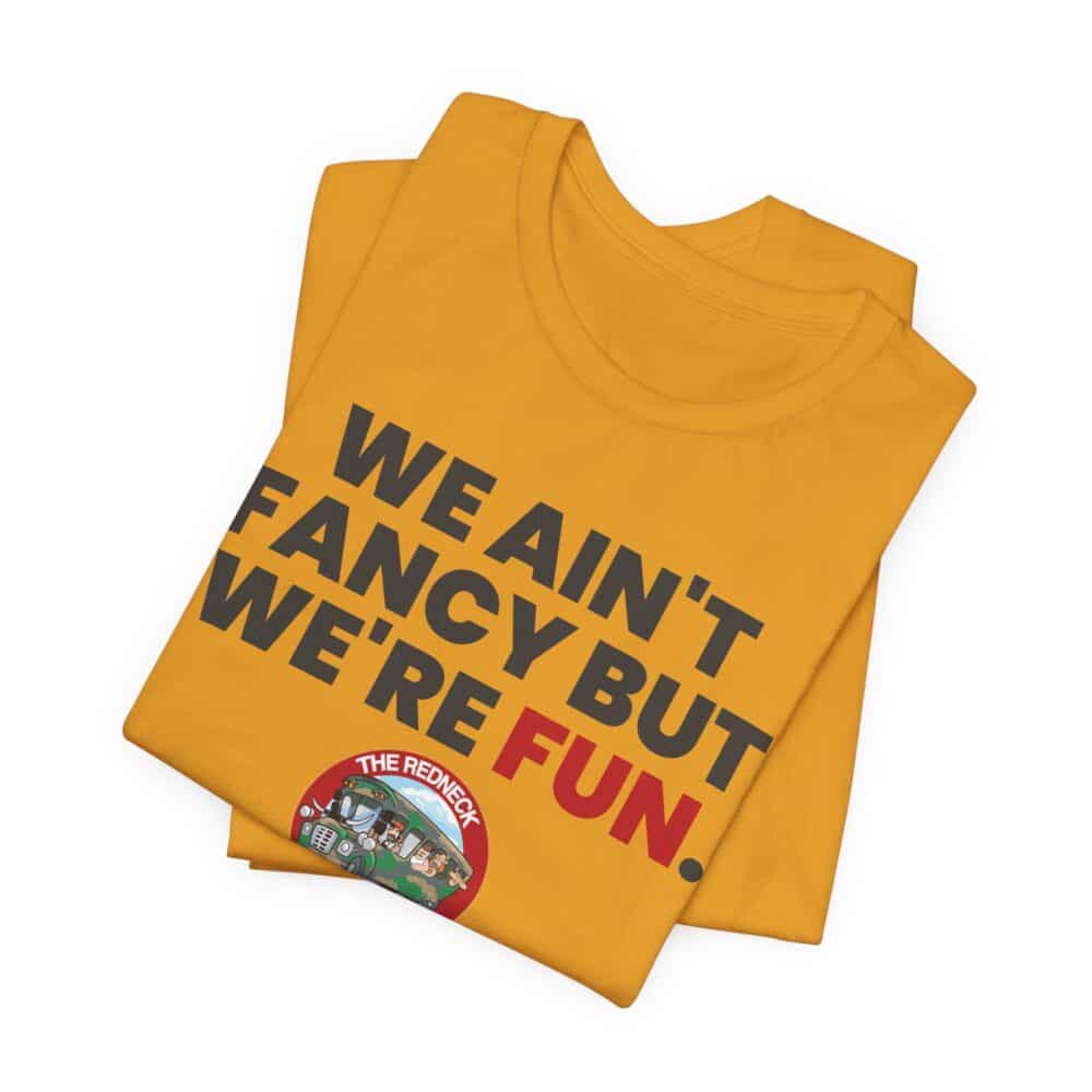 Folded We Ain't Fancy Tshirt in yellow featuring bold text "WE AIN’T FANCY BUT WE’RE FUN." and a small illustrated "THE REDNECK" logo.