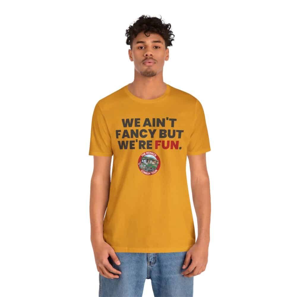 A person models the We Ain't Fancy Tshirt in mustard yellow, featuring "WE AIN'T FANCY BUT WE'RE FUN." and a small round logo beneath the text, against a plain white background.