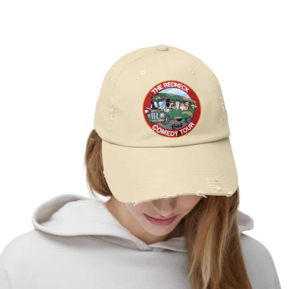 A person in a white hoodie looks down while wearing the Redneck Comedy Tour Distressed Hat, a light tan baseball cap with a cartoon car patch.