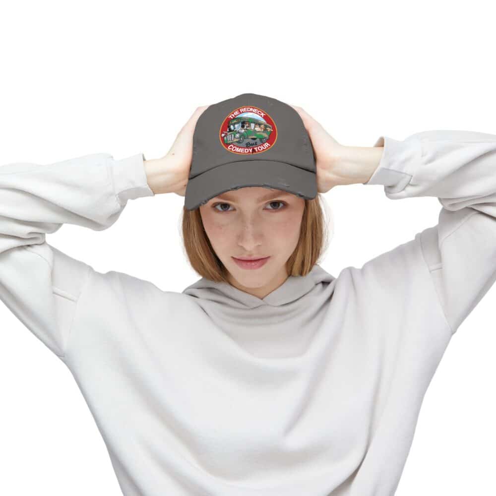 Wearing a light gray hoodie, a person holds the brim of the Redneck Comedy Tour Distressed Hat, which features a colorful "The Official Comedy Tour" embroidered patch.