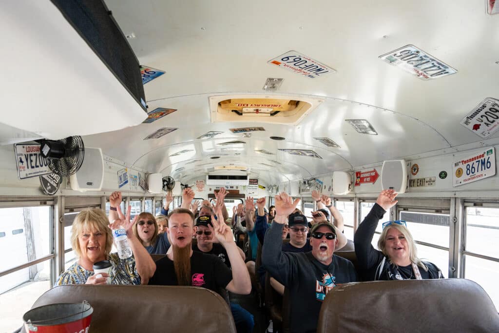 A group of people sit inside a decorated bus, smiling and raising their hands in excitement, with license plates attached to the ceiling and walls.