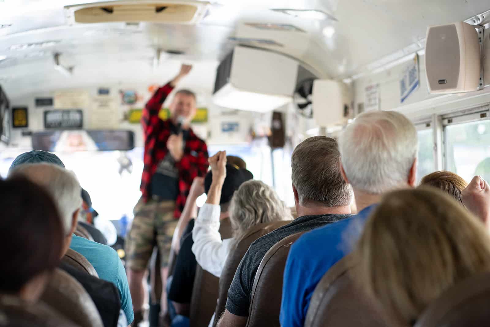 A group of people sit on a bus while a person at the front, wearing a red plaid shirt, gestures and speaks to the passengers.