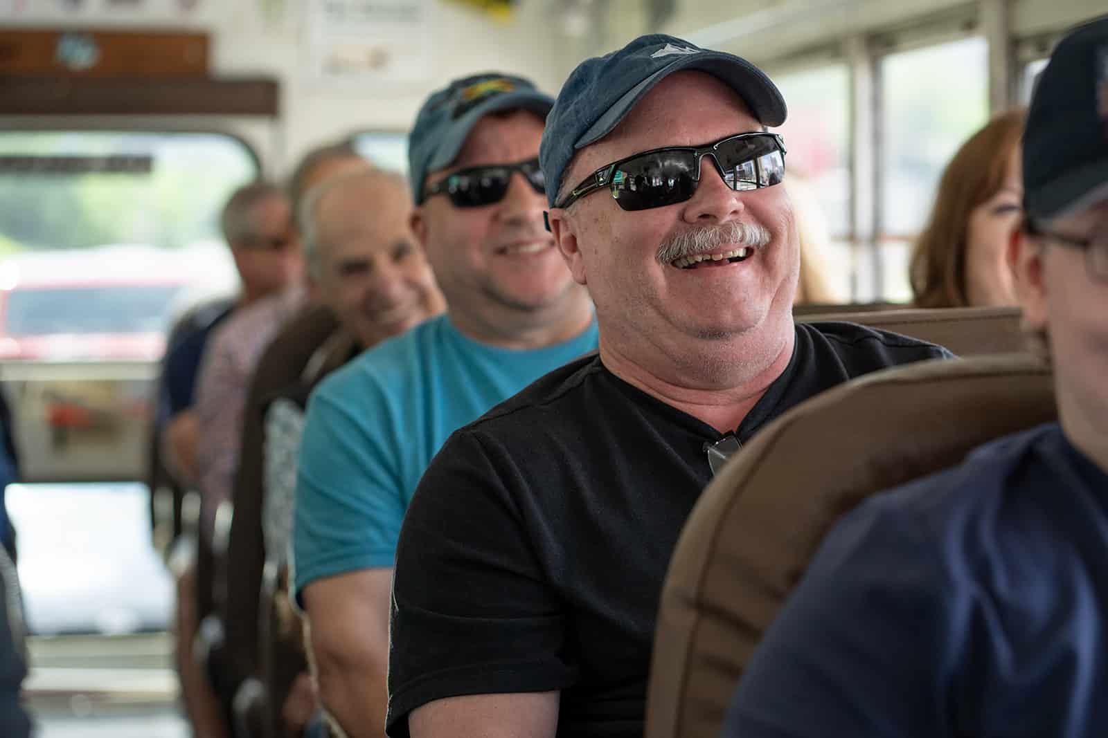 Several people, mostly men wearing sunglasses and casual clothing, sit smiling and laughing on a bus during daylight.