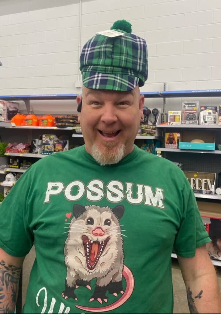 A smiling man wearing a green plaid hat and a green "POSSUM" T-shirt stands in a store aisle with shelves of various products behind him.