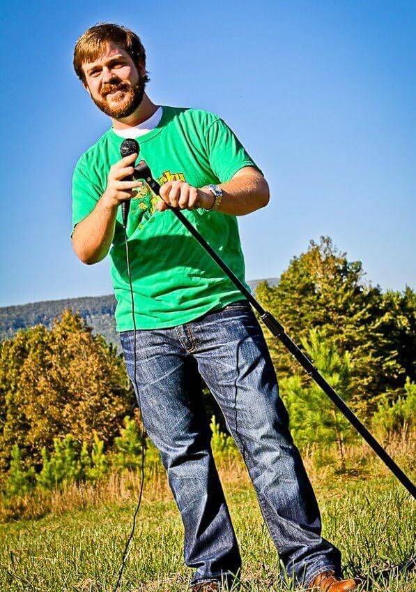 Man in a green T-shirt and jeans holding a microphone stands outdoors on grass, with trees and a blue sky in the background.