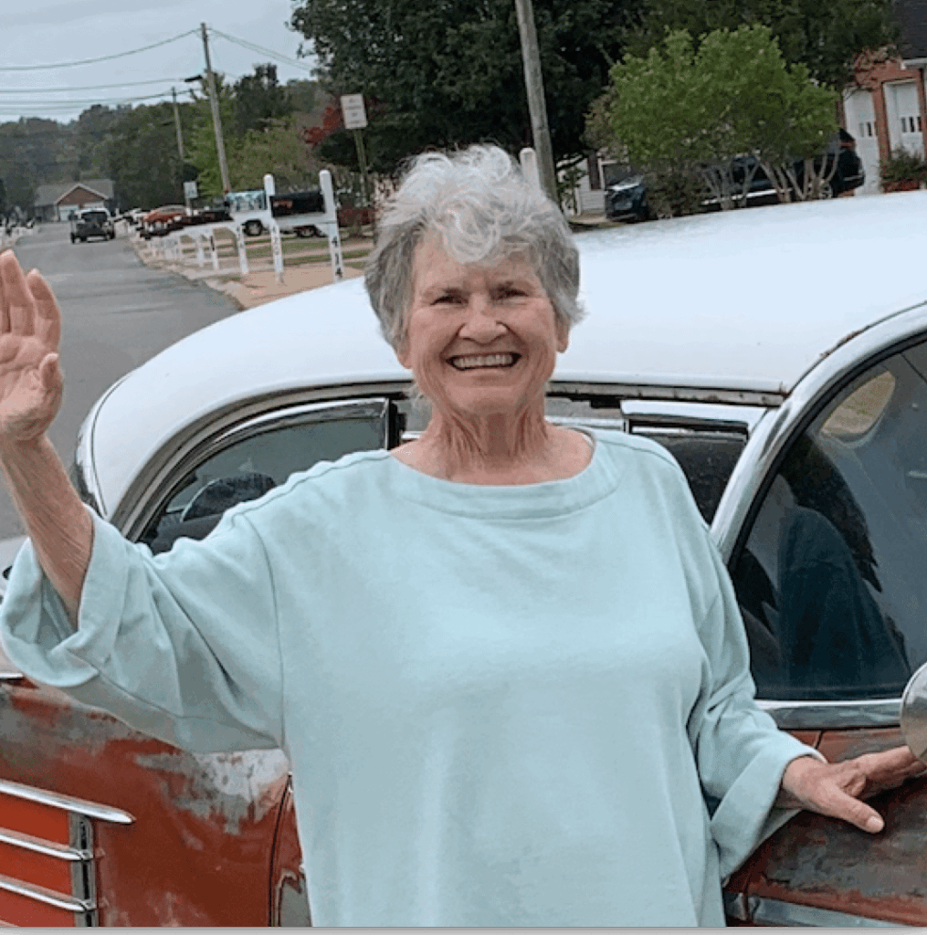 An older woman with gray hair, wearing a light blue top, stands outdoors beside a vintage car and smiles while waving.