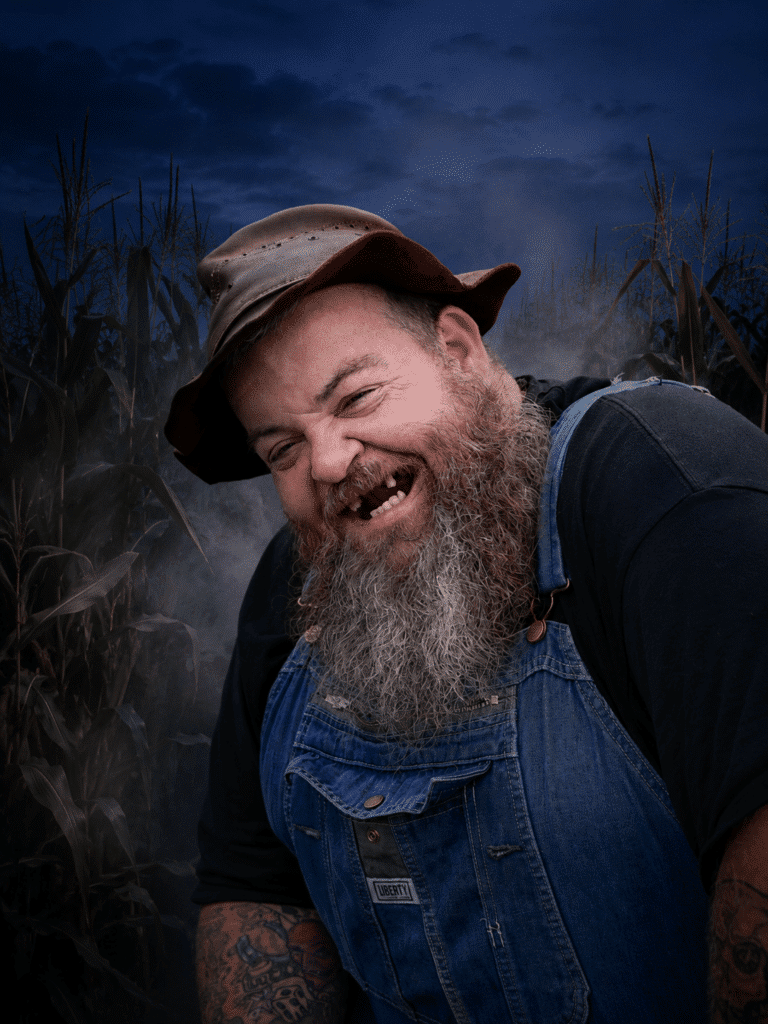 Bearded man in overalls and a hat smiles broadly, showing missing teeth, standing in front of tall corn plants at dusk.