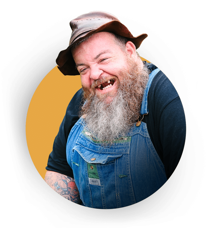 Man with a long gray beard, missing teeth, wearing a brown hat, black shirt, and denim overalls, smiling against a yellow background.