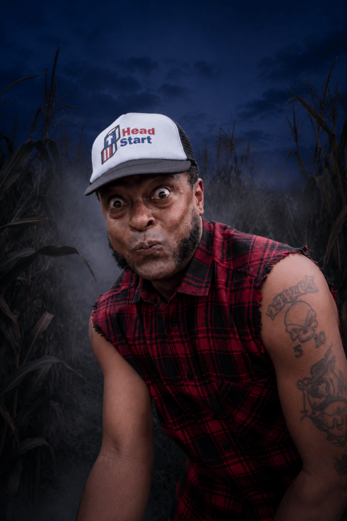 A man in a sleeveless plaid shirt and "Head Start" cap stares intensely, standing in a cornfield at dusk. Tattoos are visible on his arm.