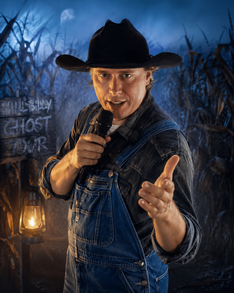 A person in overalls and a cowboy hat holds a microphone, gesturing forward, with a “Hillbilly Ghost Tour” sign and cornfield backdrop at night.